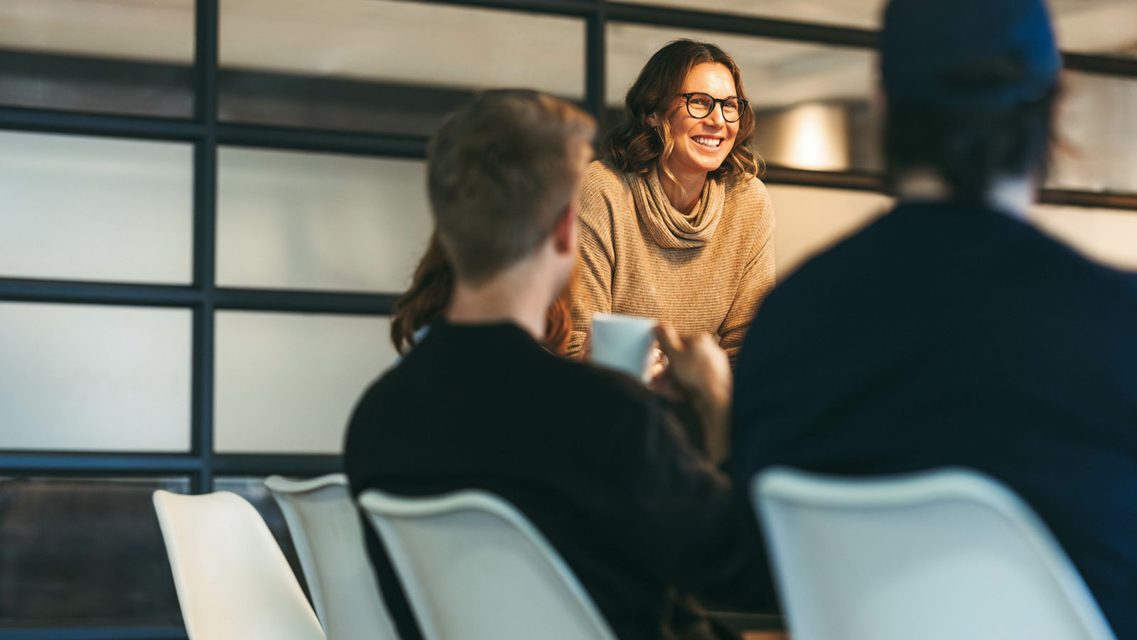 In a bright office, a diverse group of colleagues engages in a dynamic discussion. A confident woman leads the team, presenting ideas to attentive businessmen and businesswomen. Their collaboration and creativity drive their tech startup's success.