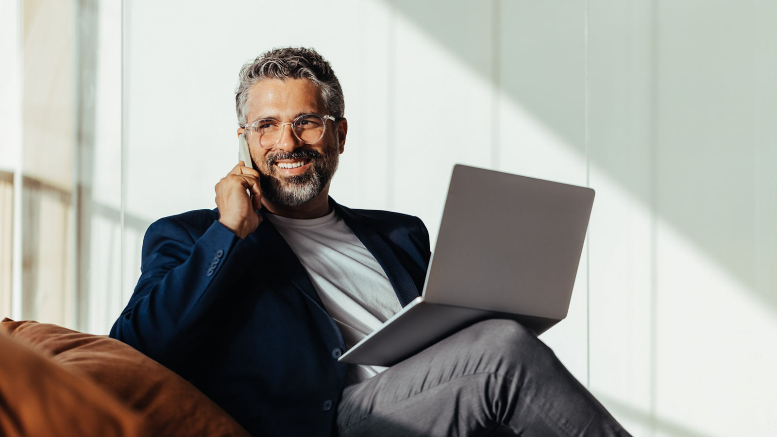 Happy, mature businessman talking on his phone while holding his laptop, is comfortably seated in a modern office. Senior entrepreneur engaging in business communications.