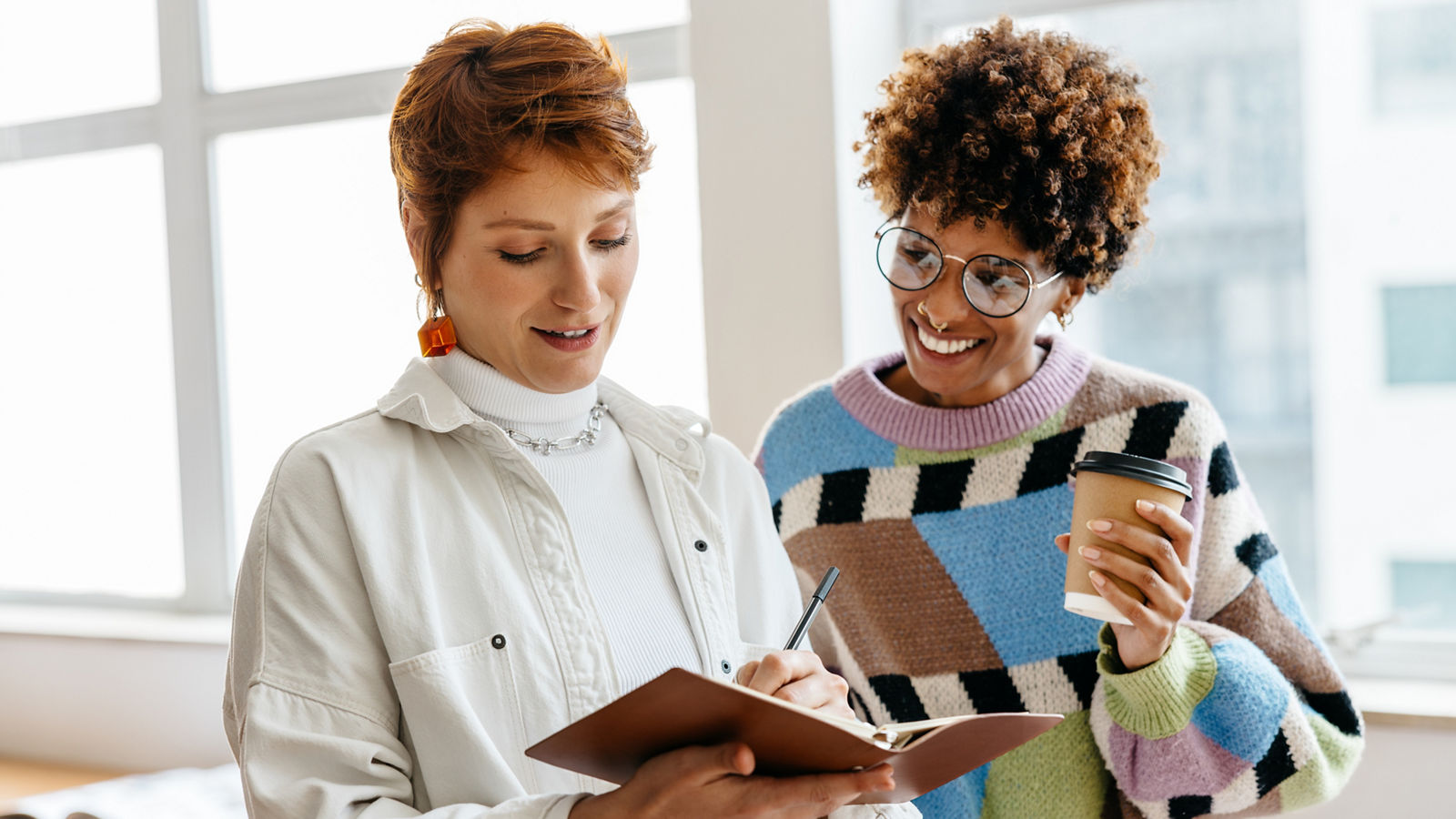 Two colleagues are engaged in a collaborative work discussion in a well-lit freelance office setting. They share ideas and notes in a co-working space, exemplifying teamwork and professionalism in a casual business environment.