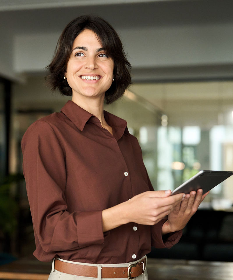 Happy young Hispanic professional business woman using tab standing in office. Happy businesswoman manager female executive leader holding tab at work looking away dreaming of future successful career