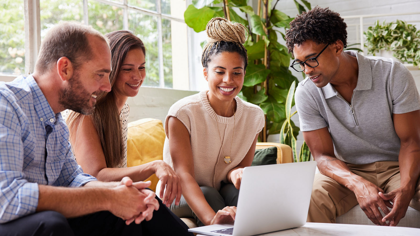 Smiling group of diverse young businesspeople talking over a laptop during a casual meeting in the lounge area of a modern office