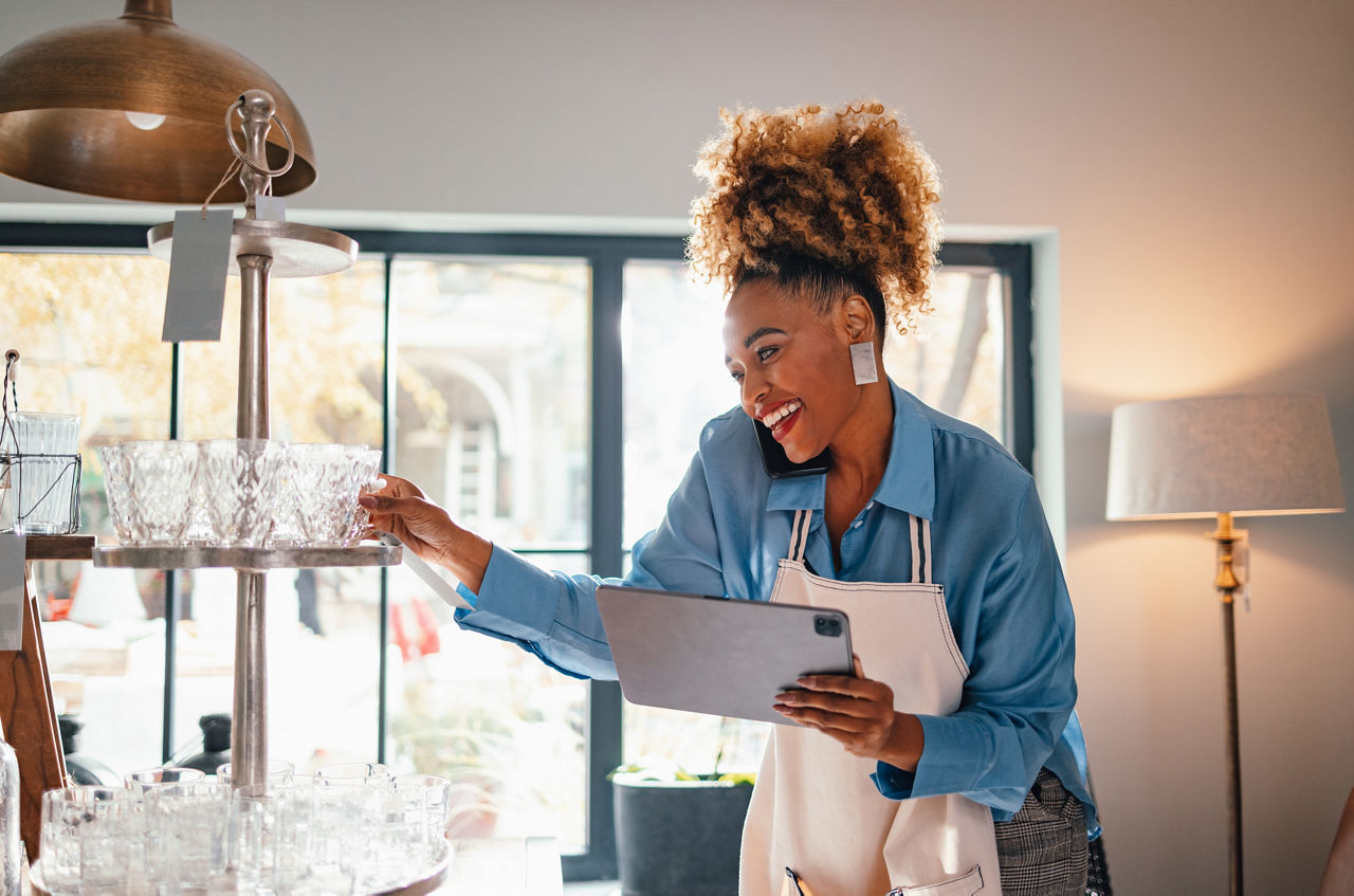 Happy businesswoman in apron holding a digital tablet while speaking on the phone with a client and standing in the store.