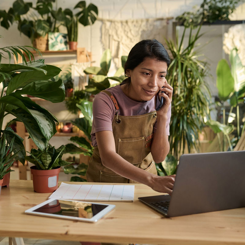 Smiling houseplant store employee talking on smartphone and checking laptop standing behind counter