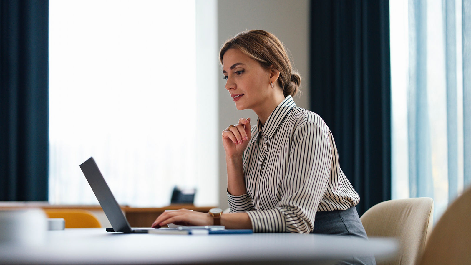 Focused professional woman with a confident look working on a laptop at an office desk, embodying productivity and dedication.