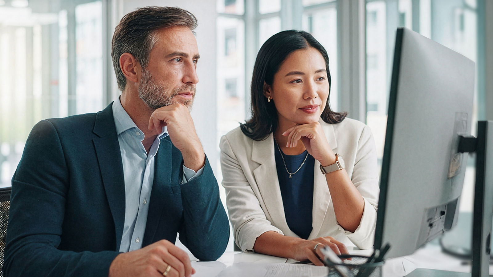 Man and woman in business attire sitting at computer, in glass office