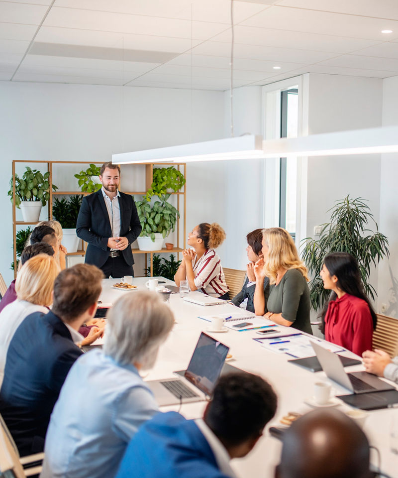 Wide angle view of business people sitting side by side at board room conference table and listening to young male colleague give presentation.