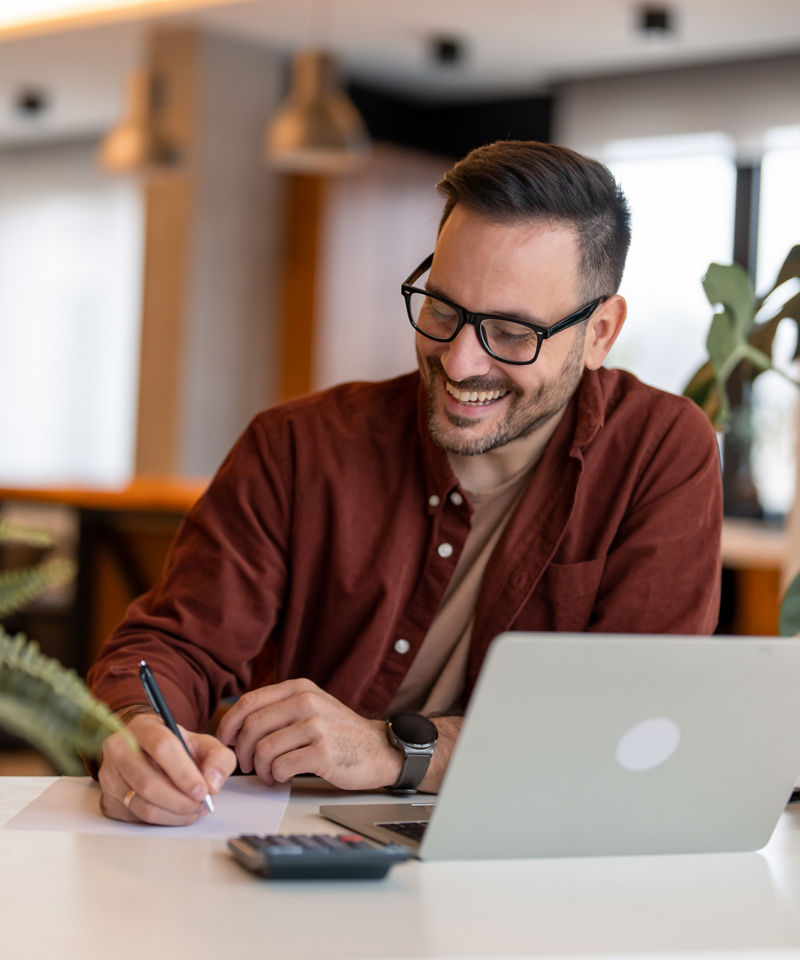 Shot of a young businessman using a laptop and writing on a paper.