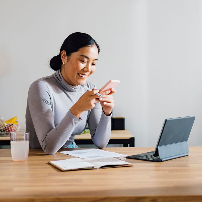 Businesswoman using digital tablet and notebook while typing text message on her mobile phone at kitchen desk.