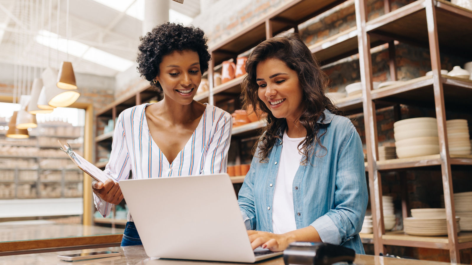 Happy young ceramists using a laptop while working in their store. Female entrepreneurs managing online orders on their website. Cheerful young businesswomen running a creative small business together.