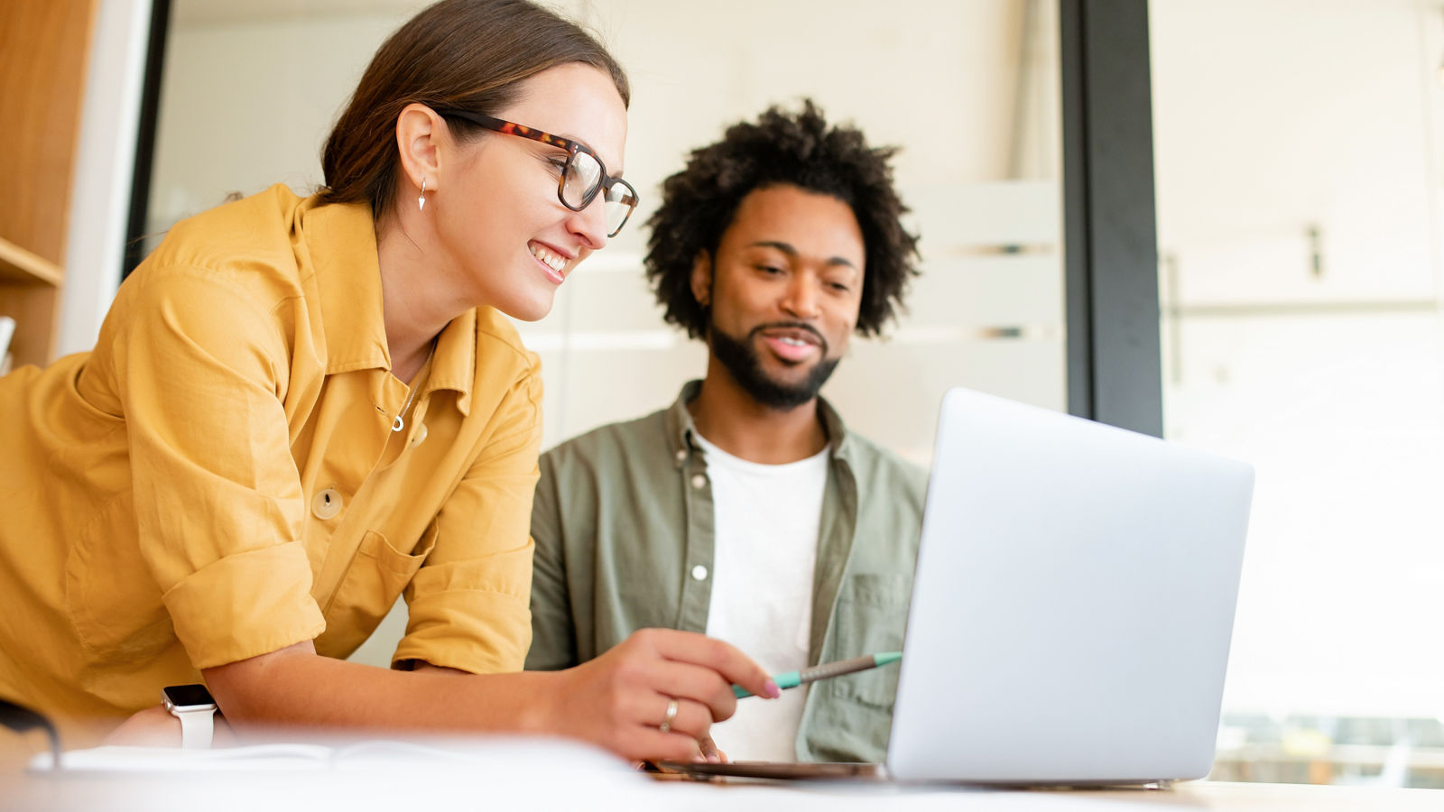 Two colleagues have pleasant conversation sitting side by side in modern open space office in front of laptop, smiling and enjoying friendly working atmosphere, brainstorming together, solving tasks