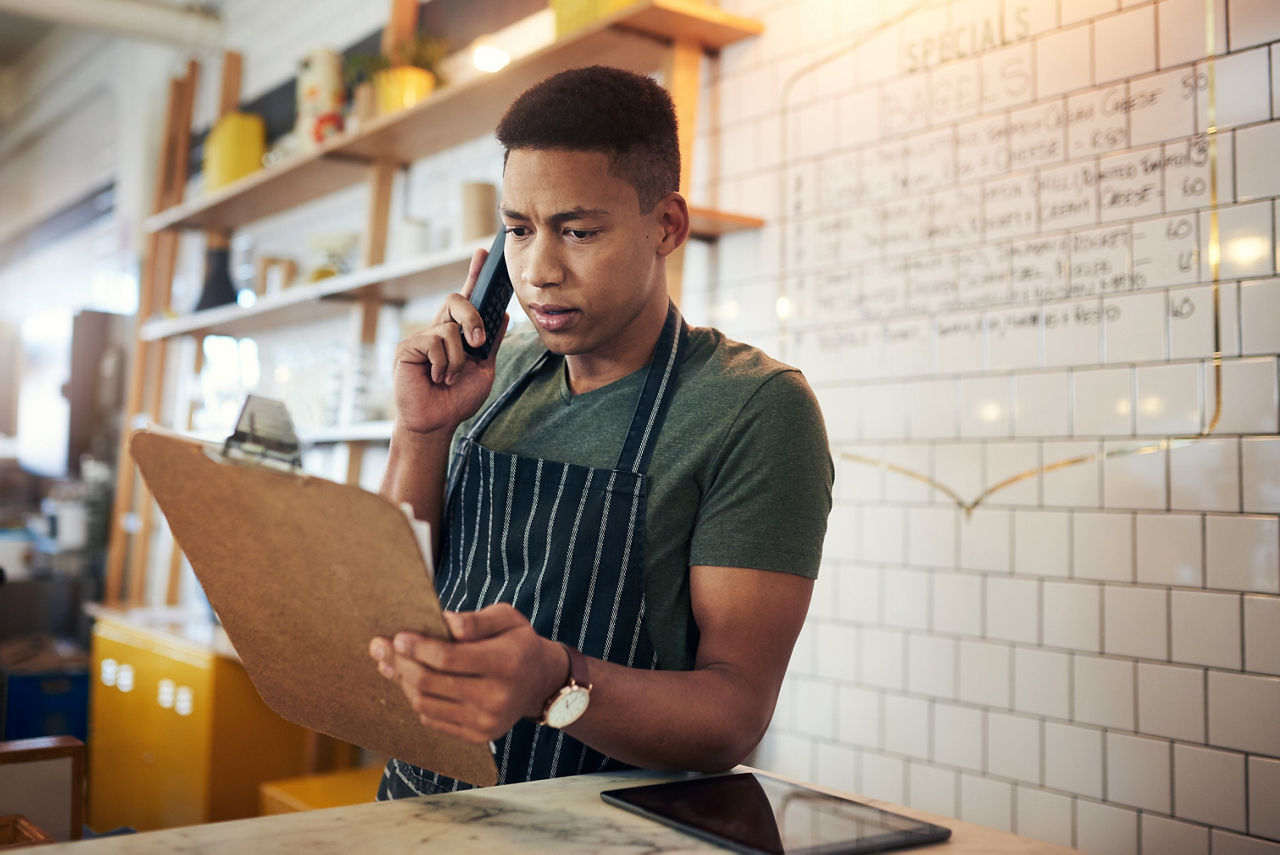 Shot of a young man talking on a phone while going through notes on a clipboard in a cafe