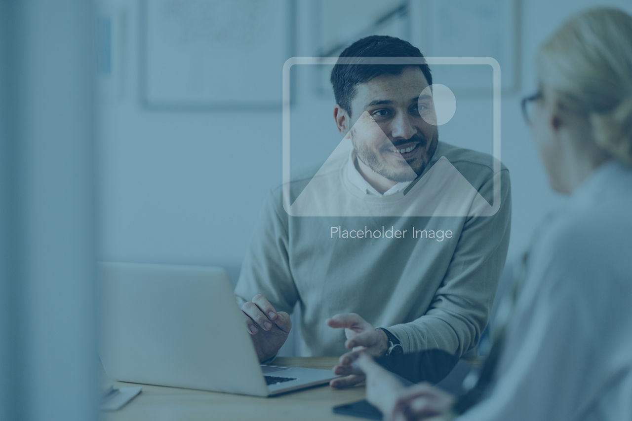 Caucasian businessman is sitting in an office and having a meeting with a female colleague. He might be working from home. It's morning or later in the day. He is smiling. A laptop is in front of him and the woman is holding a digital tablet.