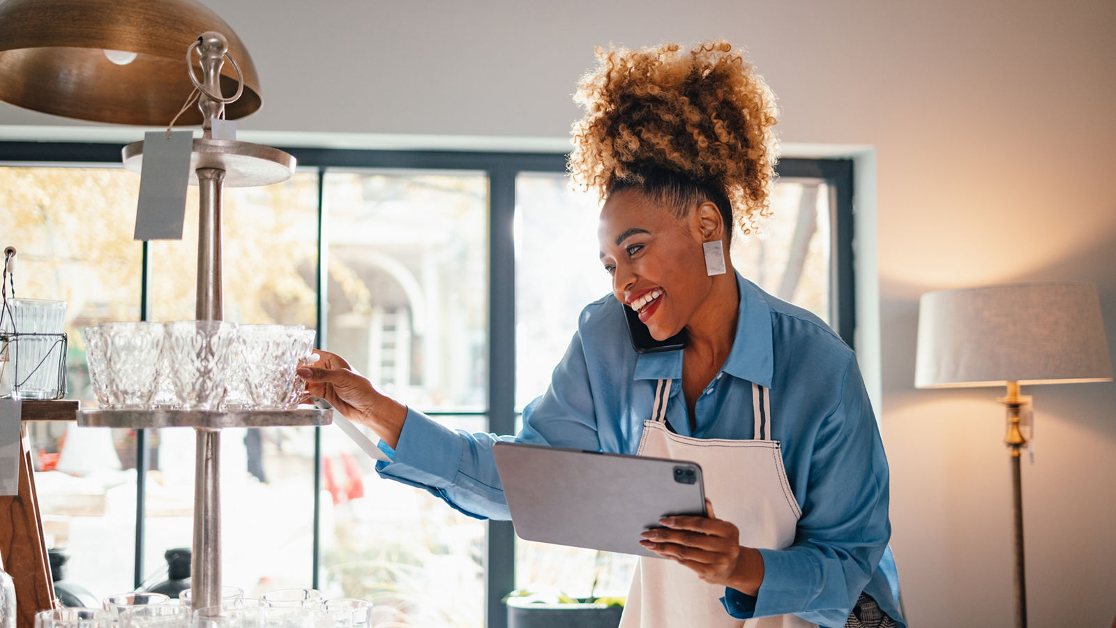 Happy businesswoman in apron holding a digital tablet while speaking on the phone with a client and standing in the store.
