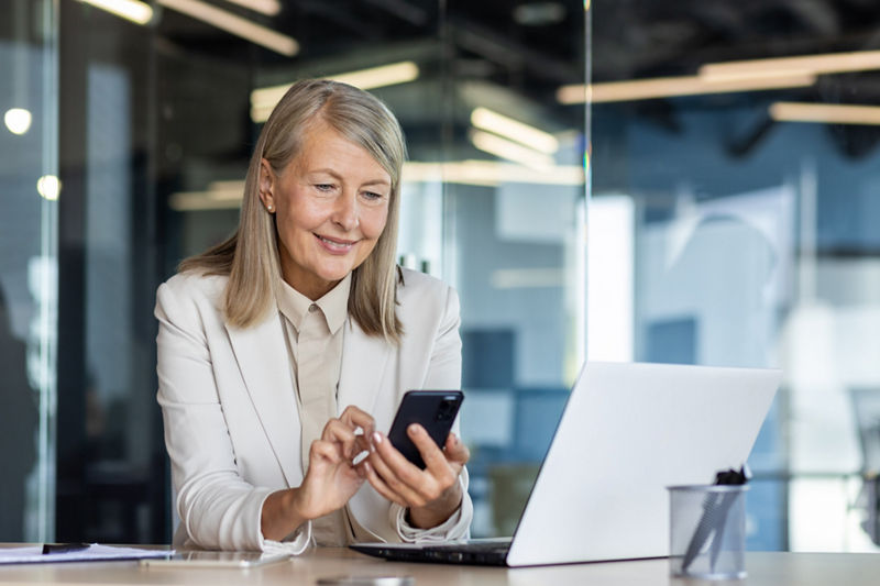 Woman at workplace inside office, business woman smiling using phone sitting with laptop, using smartphone