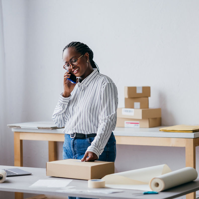 A businesswoman packing a box while talking on the phone in her home office. Concept of work from home, e-commerce, and small business.
