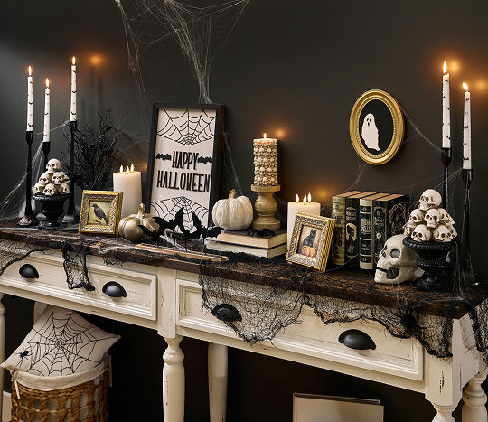A spooky Halloween-themed foyer table is decorated with classic Halloween decorations from pOpshelf featuring skeletons, Halloween skulls, moody gothic-inspired framed art, bat candlesticks, a spider web tablecloth, brass pumpkins, and more affordable Halloween themed finds.