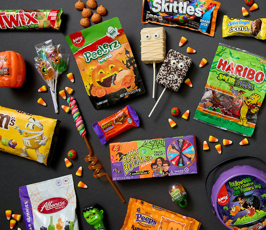 Novelty Halloween treats and candy from pOpshelf including M&Ms, Peelerz, jelly beans, Twix, candy corn, Haribo and more are displayed on black flooring.