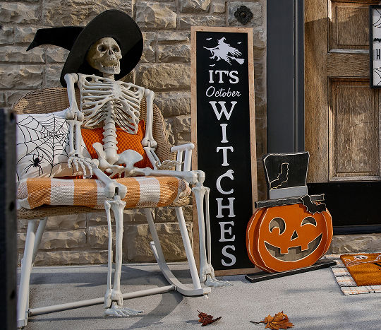 A stone home’s front porch is decorated for Halloween with affordable Halloween decor from pOpshelf, featuring full-sized decorative skeletons, a decorative candy cauldron, various pumpkin accessories, and a front porch leaner that says “It’s October Witches.”