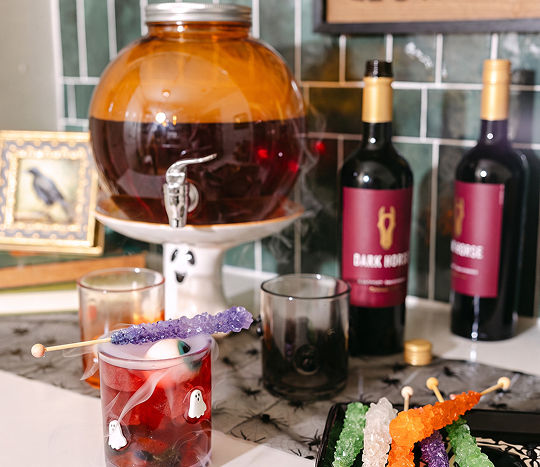 A wet bar with emerald green tile displays an orange Halloween drink dispenser and is decorated with various Halloween accents from pOpshelf including a spider web table runner, spooky Halloween icon glasses, and seasonal candies.