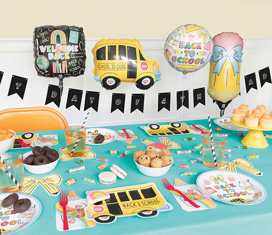 A classroom table is covered with a back-to-school themed table cover and set with affordable snacks, decor, paper plates, and balloons from pOpshelf.