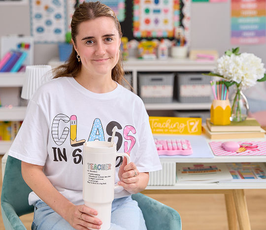 A teacher is sitting at her desk smiling at the camera while holding a teacher mug in a classroom decorated with cute affordable classroom decorations including bulletin boarders, classroom posters for teachers, and aesthetic desk decor from pOpshelf.