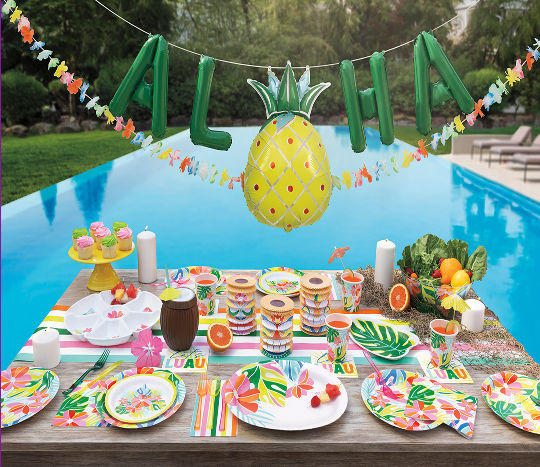 A tropical themed party tablescape decorated with colorful decorations and fruit accents next to an outdoor pool.