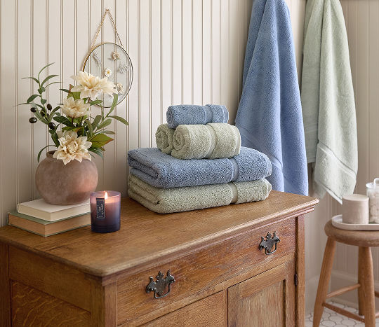 A small table is decorated with neutral toned accents featuring a vase with flowers stacked on top of storage books and accented with a candle next to a stack of neatly folded bath towels in blue and green. 