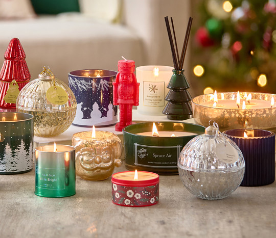 Christmas candles in various sizes, designs, and scents on a table in front of a Christmas tree.