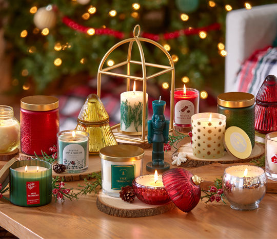 Christmas candles in various sizes, designs, and scents on a table in front of a Christmas tree.