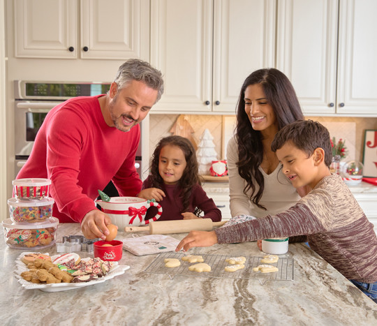 Family baking in kitchen with Christmas mixing bowl, cookie sheets, cookie cutters, tree-shaped ceramic platter, plastic food storage and more in a kitchen decorated for Christmas.