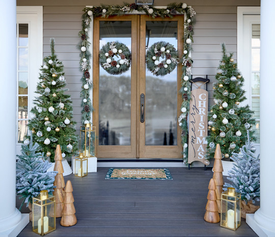 Front porch decorated for Christmas with 6-ft lighted trees, 3-ft. potted trees with fairy lights, sled porch leaner, wooden trees, garland & wreaths, and gold lanterns.