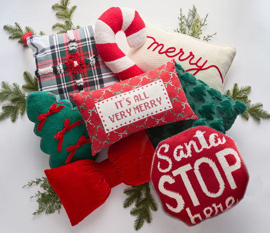 Christmas pillows in various shapes and designs on a sofa: green tree with bows, candy cane, giant red bow, plaid, Santa Stop Here, and more.