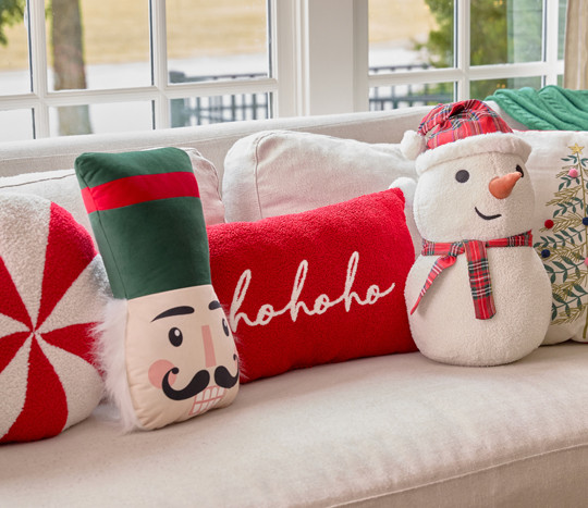 Christmas pillows in various shaped and designs on a sofa: snow globe, peppermint candy, nutcracker, snowman, red ornament & more.