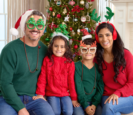 Family in front of Christmas tree wearing Santa hats, novelty Christmas glasses, Christmas bead necklaces, antler and tree headbands, and more.