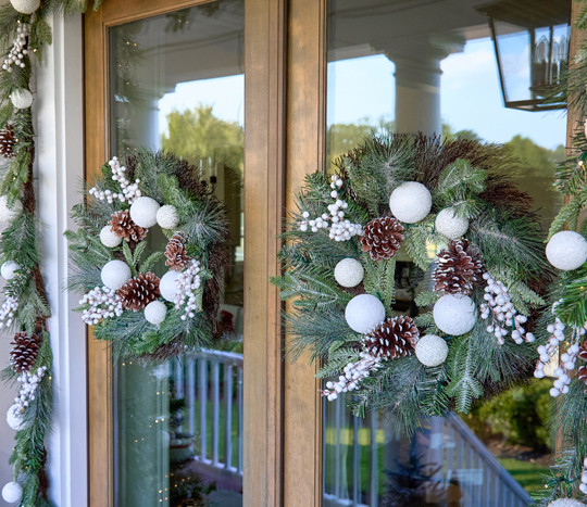 Front door with two faux floral wreaths and garland featuring white ornaments, flocked pine and pinecones, and white berries.