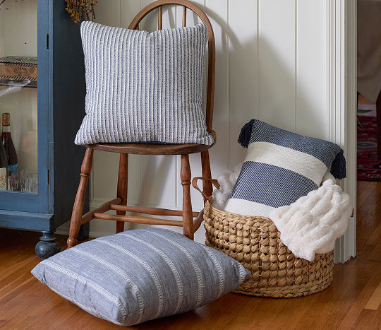A quaint wooden chair decorated with affordable pillows from pOpshelf is shown next to a basket filled with a blanket and pillow with another plush blue striped pillow shown beside the basket on the ground.