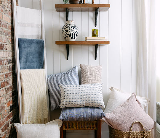 A bright living room shows a chair sitting in the corner and stacked with blue accent pillows between a throw blanket ladder display and a basket full of more affordable plush pillows.
