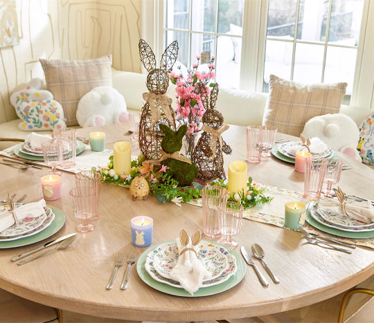 Dining table set for easter brunch with floral plates pink plastic glass and goblet, pastel green chargers and bunny ear napkin holders, with a spring floral and rattan and moss bunny centerpiece.