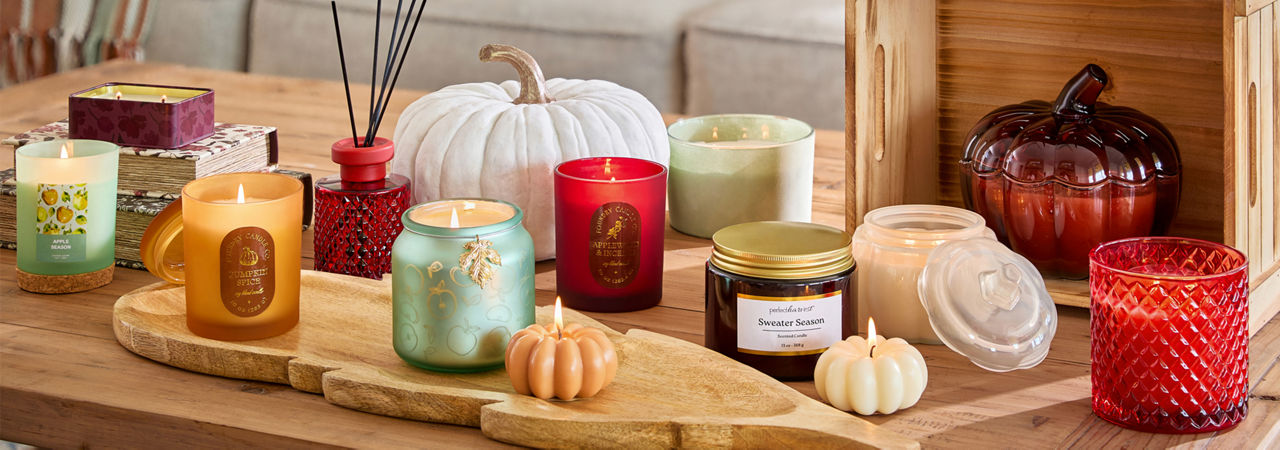 Collection of fall container and jar candles in various colors, fall scents, and shapes in front of a sofa with fall pillows.