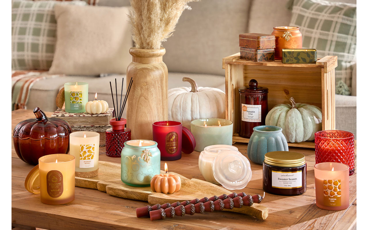 Collection of fall container and jar candles in various colors, fall scents, and shapes in front of a sofa with fall pillows.