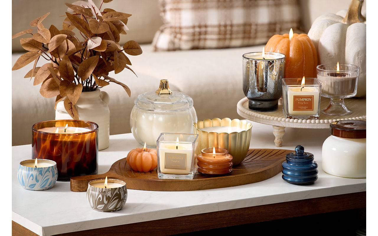 A white coffee table placed in front of  a beige couch decorated with a brown plaid pillow features various fall candles including pumpkin scents and shapes on top of a leaf shaped mango wood serving tray.