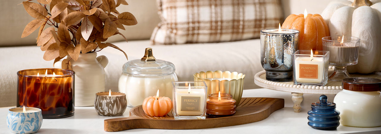 A white coffee table placed in front of  a beige couch decorated with a brown plaid pillow features various fall candles including pumpkin scents and shapes on top of a leaf shaped mango wood serving tray.