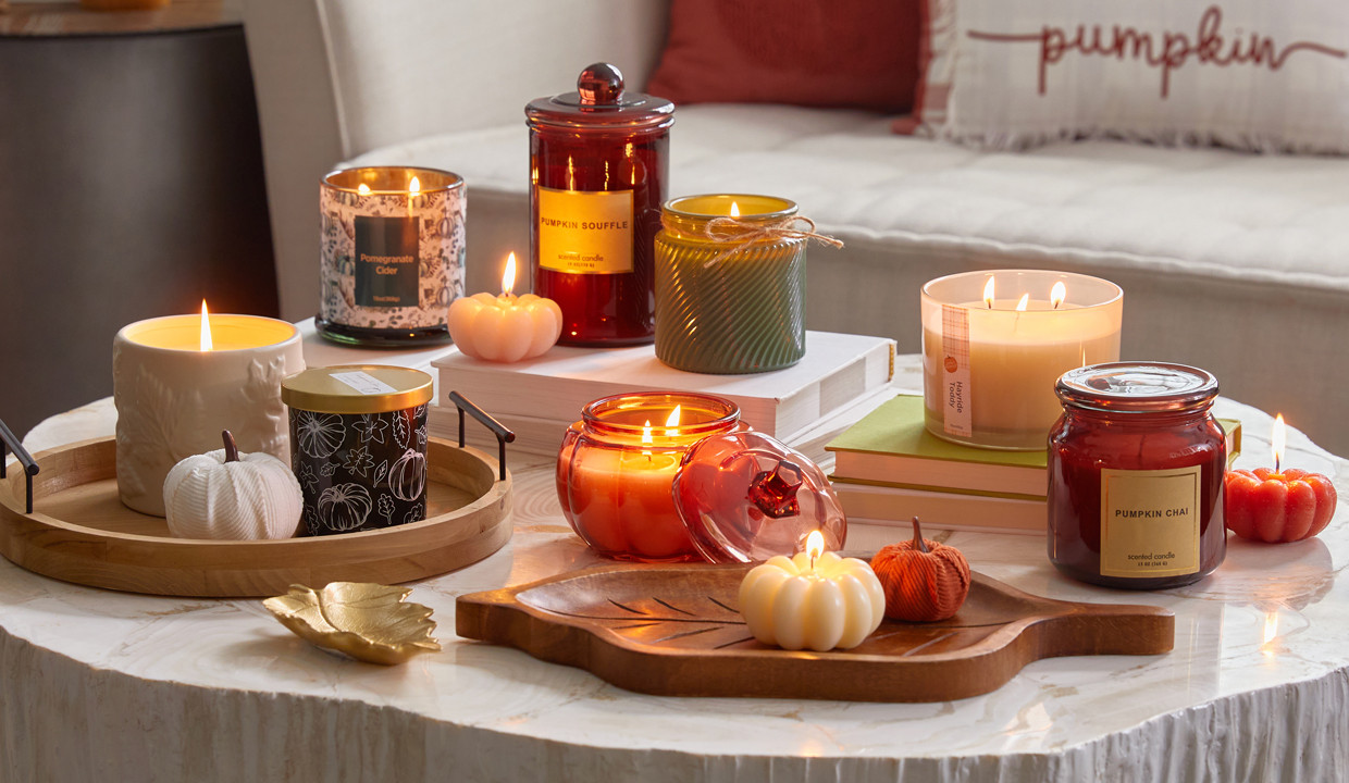 Collection of fall container and jar candles in various colors, fall scents, and shapes in front of a sofa with fall pillows.