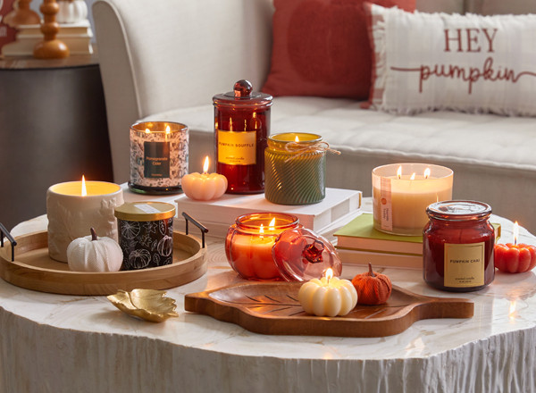 Collection of fall container and jar candles in various colors, fall scents, and shapes in front of a sofa with fall pillows.