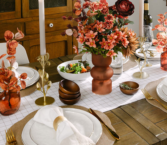 An autumn-themed dinner table features a close-up of a fall tablescape from pOpshelf in shades of orange, cream, and gold metallics with elevated dinnerware including textured plates with gold accents. 