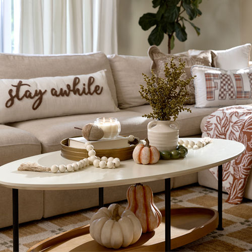 A neutral-toned living room is decorated with classic fall decor from pOpshelf in shades of orange and cream with plaid patterns, pumpkin accents, and autumn scented candles.