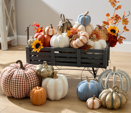 A small black wooden paneled wagon is filled with fall decor including sunflowers, autumn leaves, and various pumpkins in metallic, orange, blue & cream with more pumpkins sitting at the wheels of the wagon.