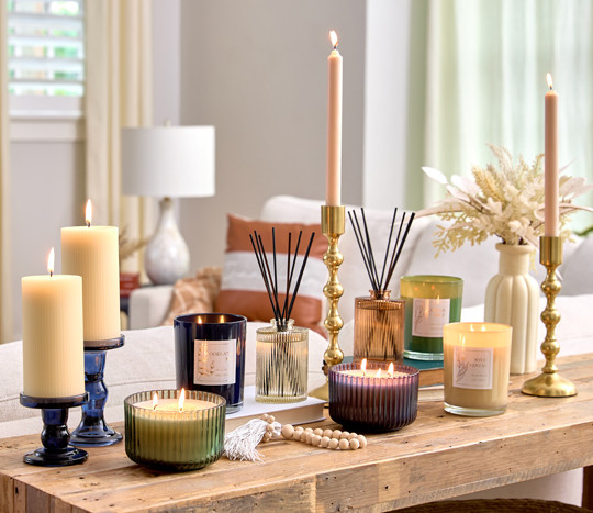 Console table set with large white pillar candles in blue glass holders, multiple reed diffusers, various jar candles, and taper candles in gold candlestick holders.