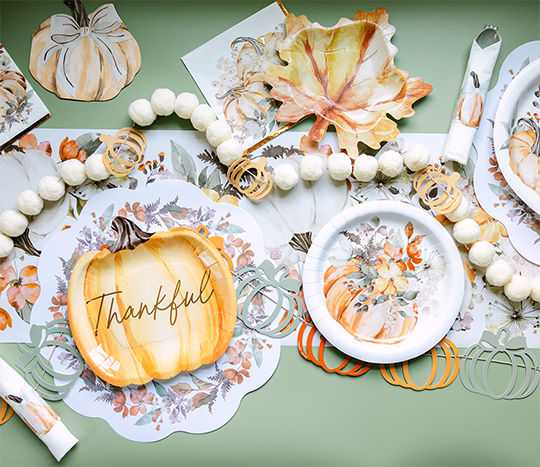 A green party table is set with a classic fall party tablescape including pumpkins, leaf shaped plates, “Thankful” pumpkin Thanksgiving themed paper plates and napkins from pOpshelf.