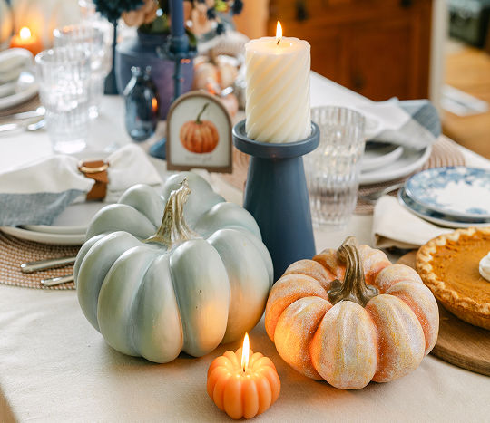 A dining table covered in a white tablecloth is decorated with fall themed decor featuring a close-up of faux pumpkins, candles, and candleholders in shades of orange, cream, and blue.